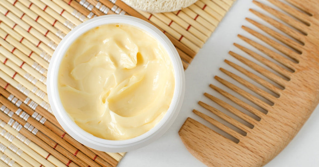 Closeup of a yellow-colored cream hair mask in a jar next to a wooden wide-toothed comb sitting on a table.