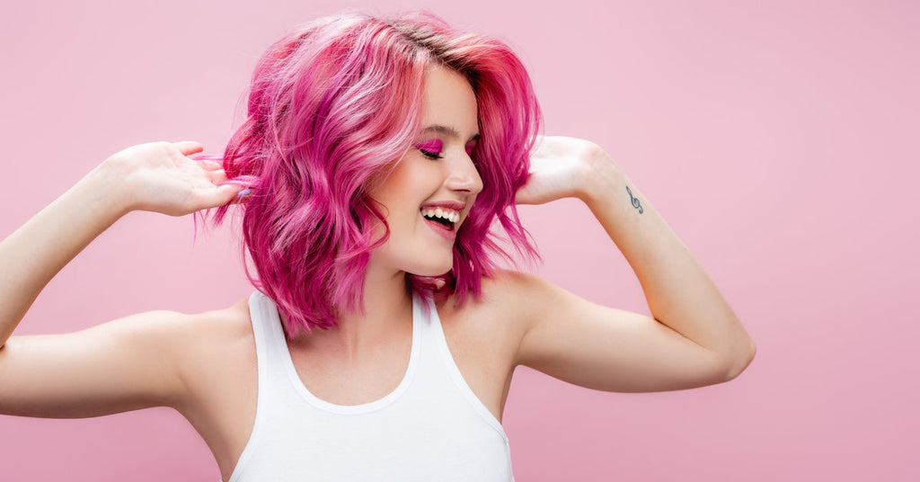 A close-up of a young woman with a short pink bob hairstyle smiling and raising her arms against a pink background.