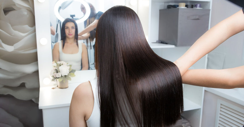 A young woman sitting in a salon chair in front of a mirror. Her treated hair is straight, shiny, and dark brown.