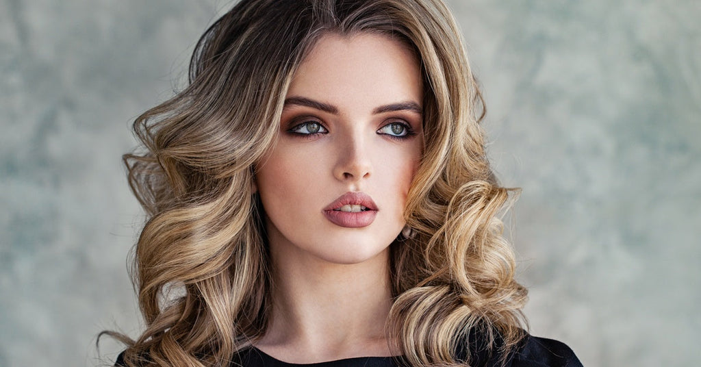 Closeup of a young, beautiful woman with a full face of makeup and voluminous curled hair against a grey background.