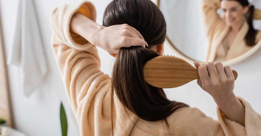 A back view of a woman in a bathrobe standing in front of a bathroom mirror while she brushes her hair into a ponytail.
