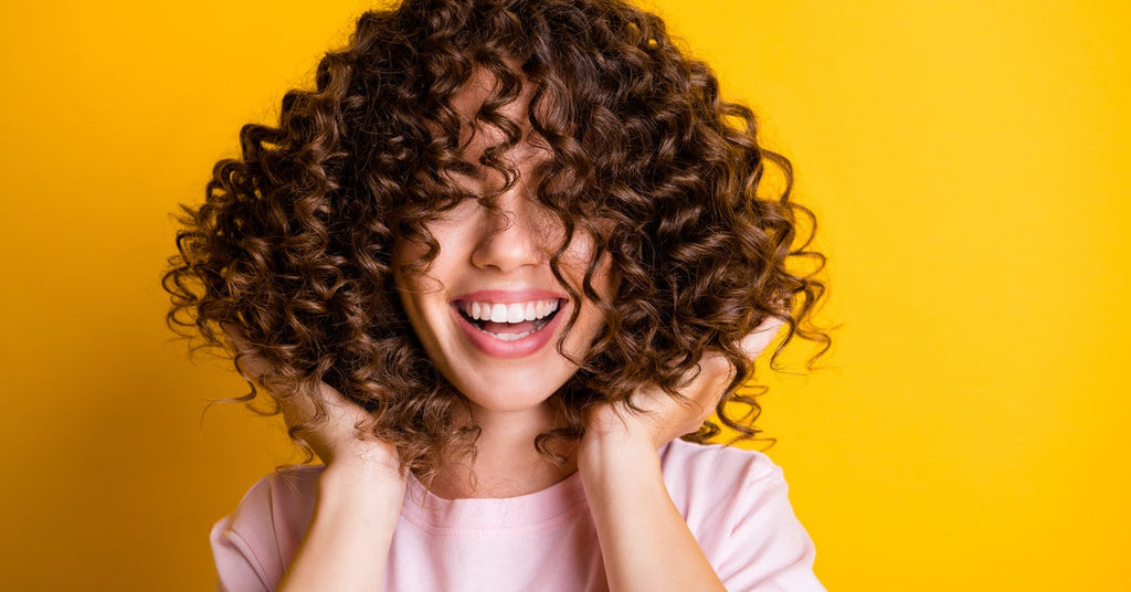 A woman in a pink t-shirt laughs as she playfully touches her curly hair against a vibrant yellow background.