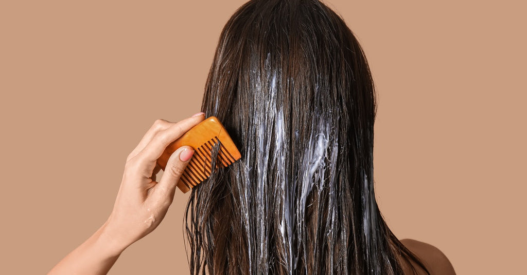 A view of a woman's back as she gently combs her long brown hair, which is covered in creamy treatment residue.