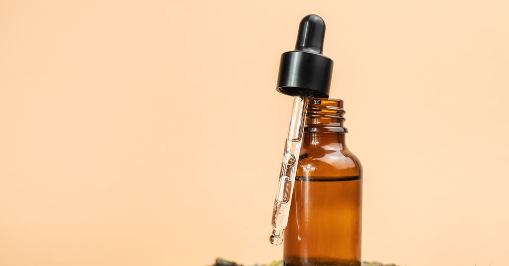 A close-up of a small glass bottle and medical pipette filled with oil in the tube resting on a bark-like surface.