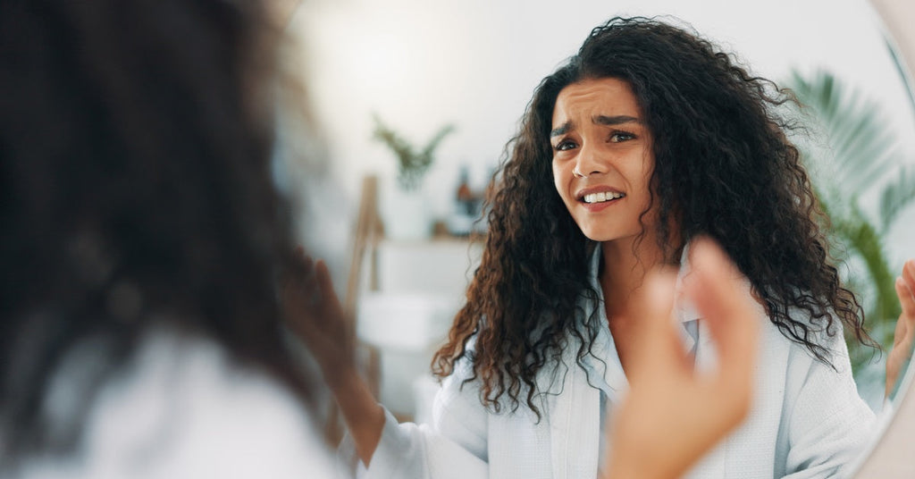 A young woman staring at herself in the bathroom mirror. She has an upset expression, and her brows furrowed.