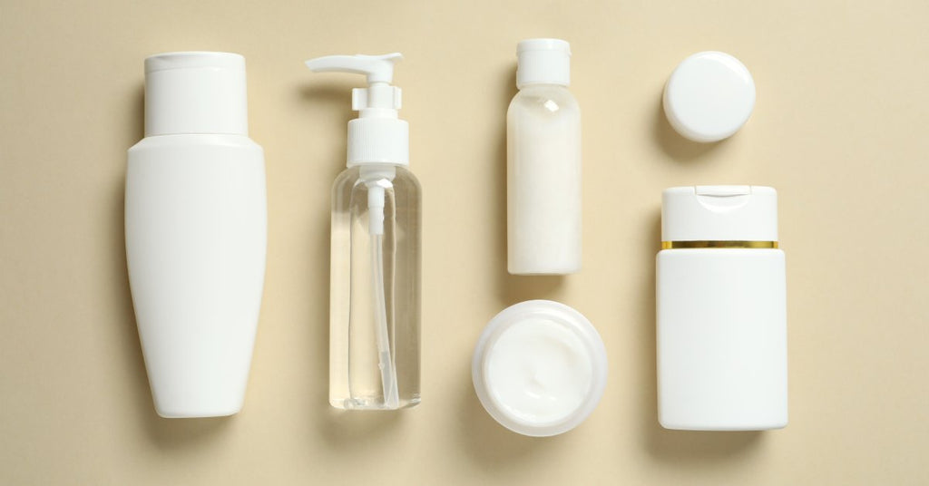 Six white travel-sized cosmetic products of varying sizes lie flat against a beige colored background.